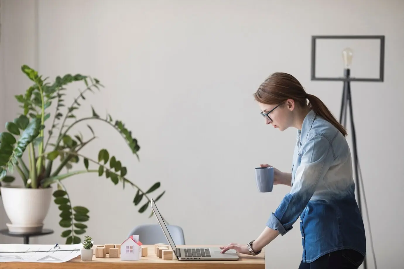 Seitenansicht einer jungen Frau, die am Arbeitsplatz am Laptop arbeitet und dabei eine Kaffeetasse hält.