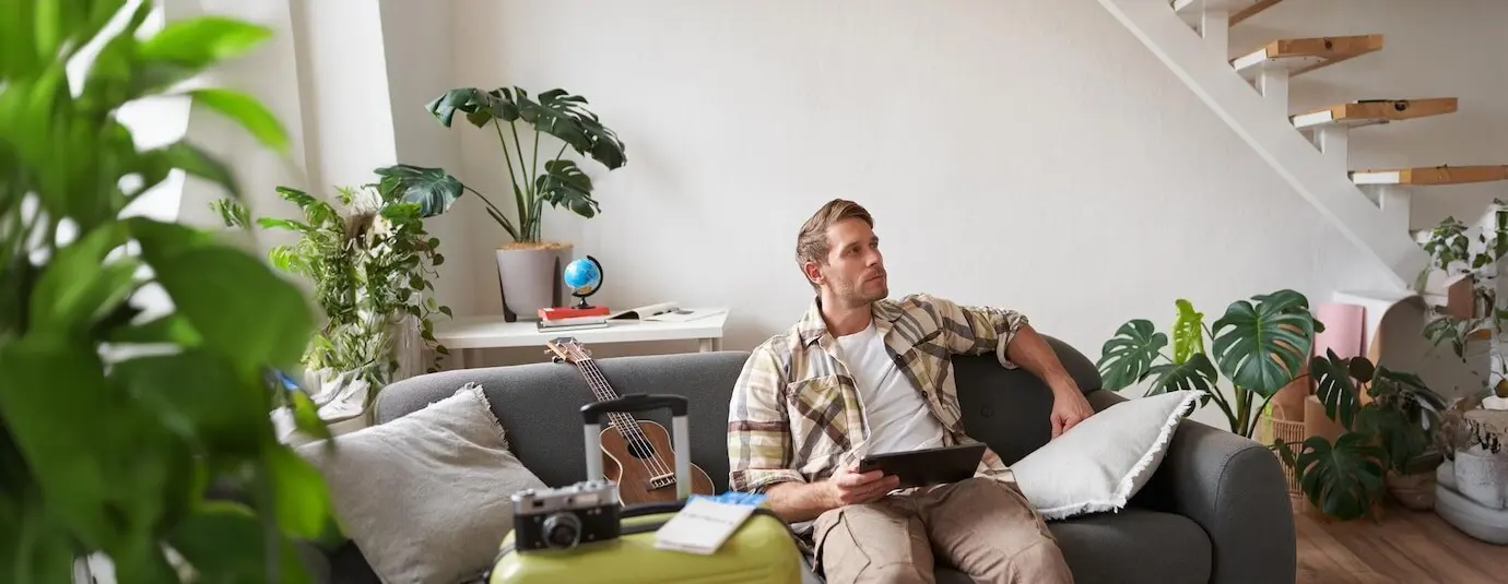 Porträt eines gutaussehenden jungen Touristen, der mit einem digitalen Tablet neben einem Koffer und zwei Tickets sitzt, online etwas für seinen Urlaub bucht und sich auf den Urlaub vorbereitet.